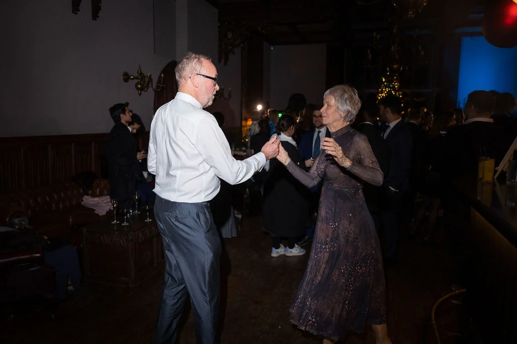 An older couple enjoys dancing together on the wooden dance floor during a lively evening wedding reception in a wood-panelled hall.