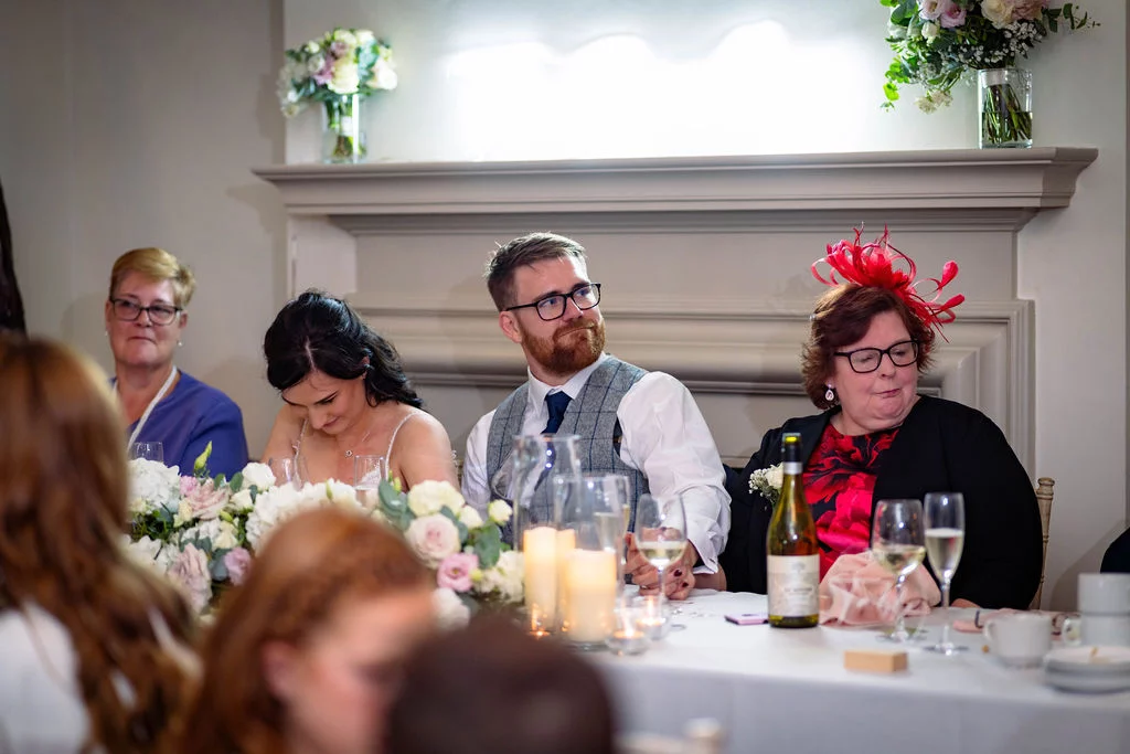 The bride and groom seated at the top table during wedding breakfast speeches, surrounded by family members, floral arrangements, and flickering candles.