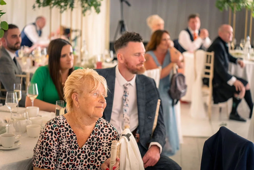 Wedding guests seated around tables during the reception, laughing and watching intently as speeches unfold in a bright, airy venue.