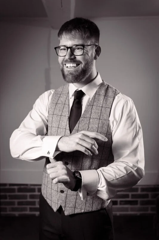 A smiling groom wearing glasses, a white shirt, a grey checked tweed waistcoat, and a tie adjusts his cufflinks in a black and white portrait.