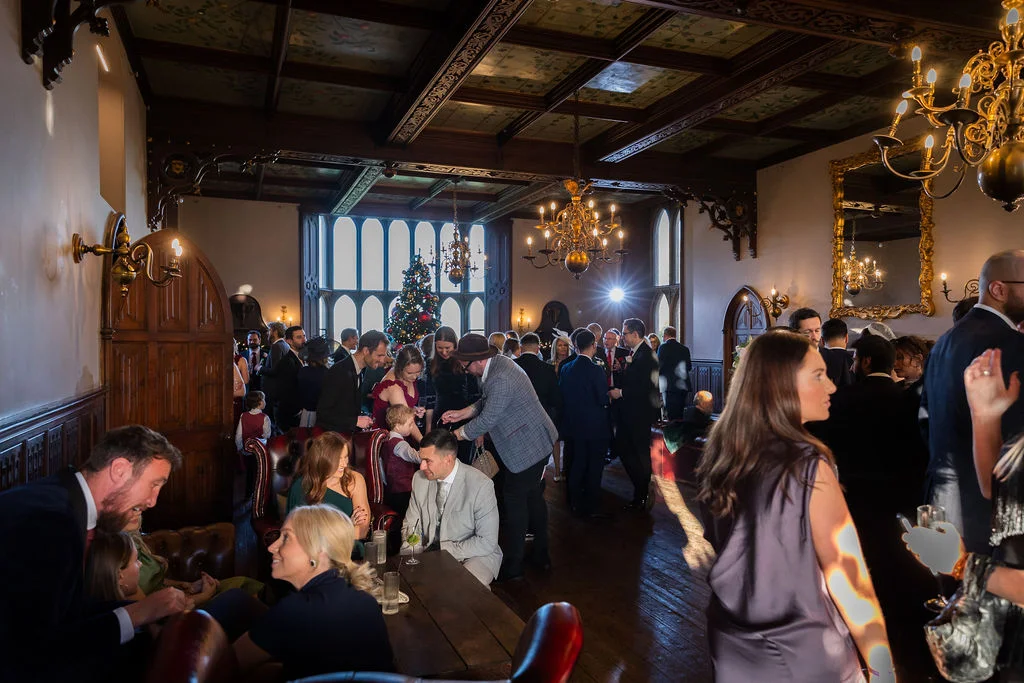 Wedding guests mingle during a lively drinks reception inside a grand, wood-panelled stately home featuring ornate chandeliers and a festive Christmas tree.