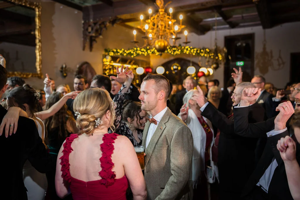 A lively evening reception inside a historic venue features guests dancing energetically beneath a grand chandelier and glowing fairy lights.