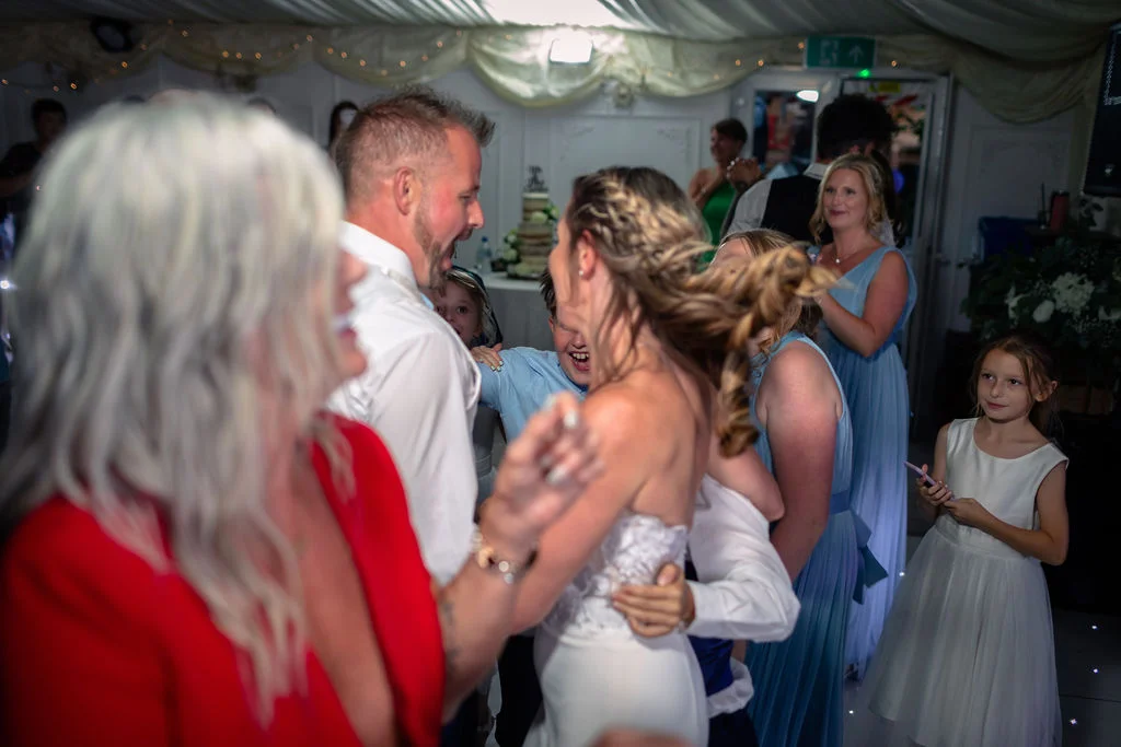 The bride and groom share an energetic first dance surrounded by family and friends in a brightly lit marquee.