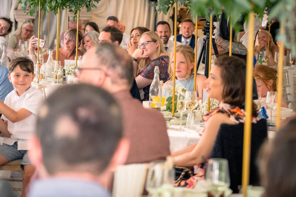 Wedding guests smiling and listening during speeches at a long banquet table decorated with tall greenery centrepieces inside a marquee.