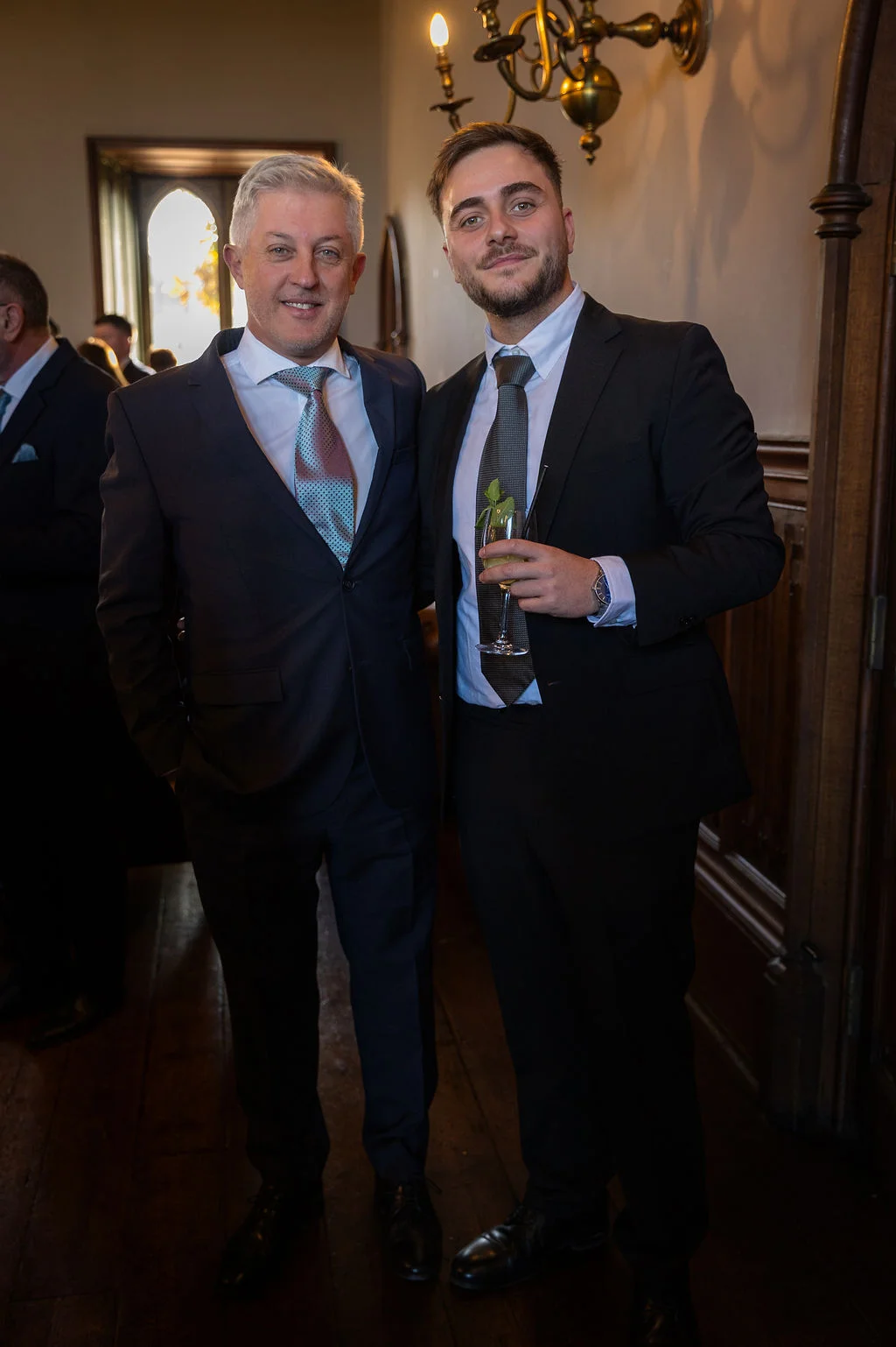 Two well-dressed male guests pose together inside an elegant wood-panelled reception room, with one holding a garnished cocktail glass.