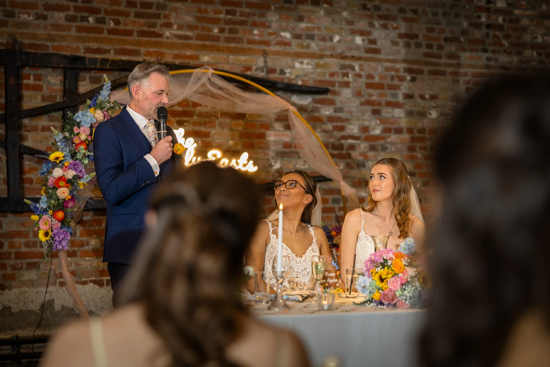 A father delivering a speech at the top table in a rustic barn venue with exposed brick walls, as two smiling brides look up at him.