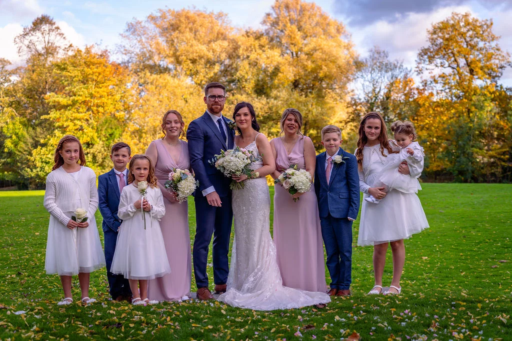 A bridal party including bride, groom, bridesmaids, and young flower girls poses outdoors on a grassy lawn surrounded by vibrant autumn foliage.