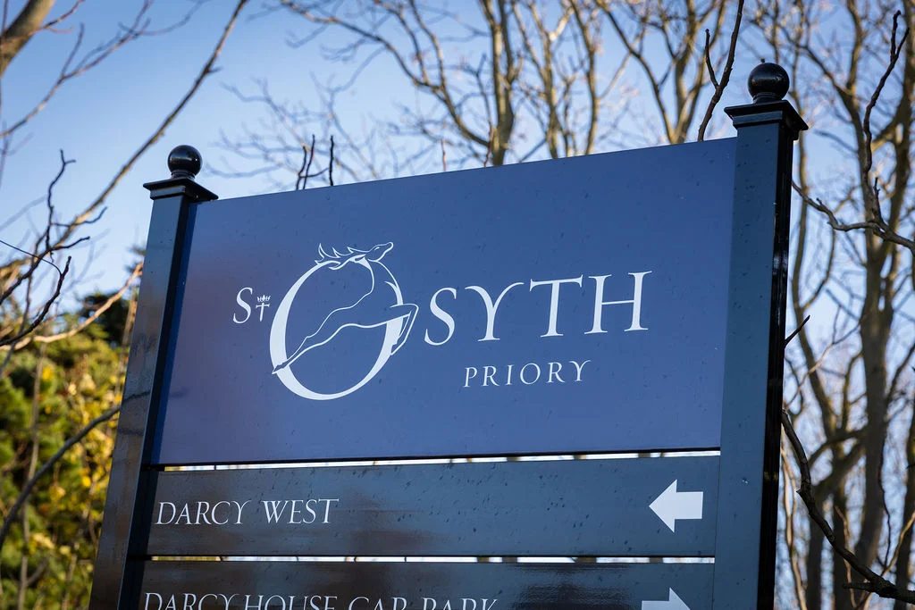 A clear blue sky frames the elegant blue and white signage for St Osyth Priory wedding venue.