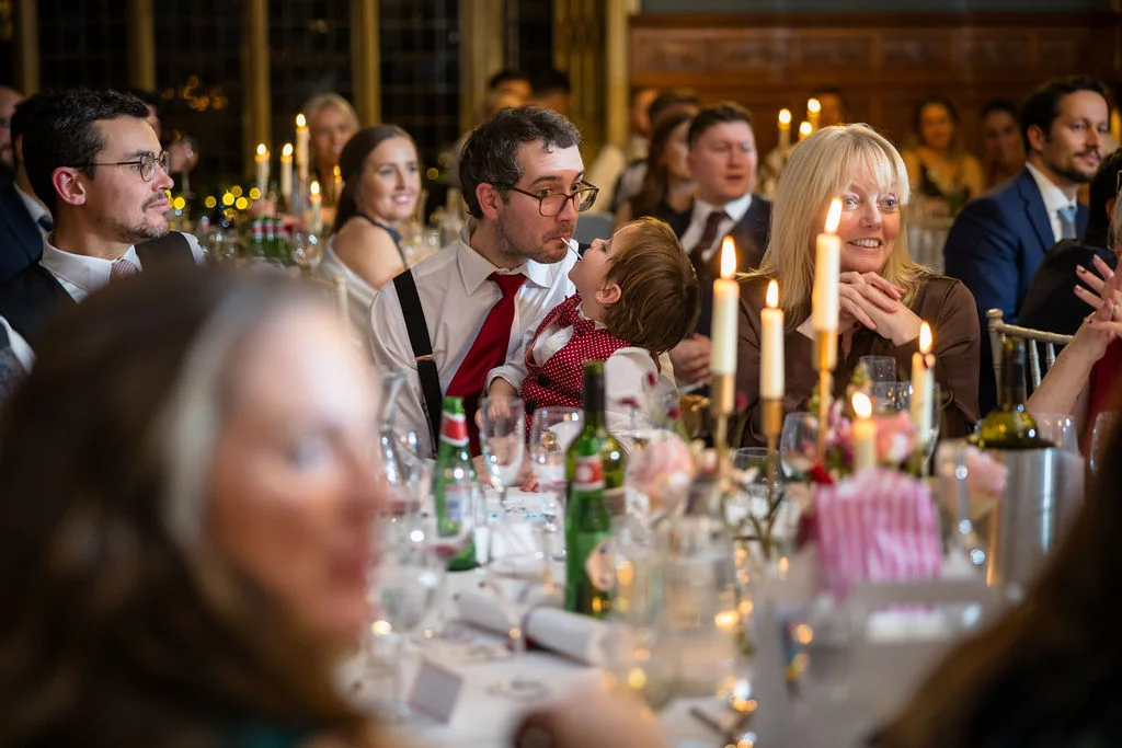 A candid moment captures wedding guests enjoying the wedding breakfast at a candlelit table, with a father interacting playfully with a toddler.