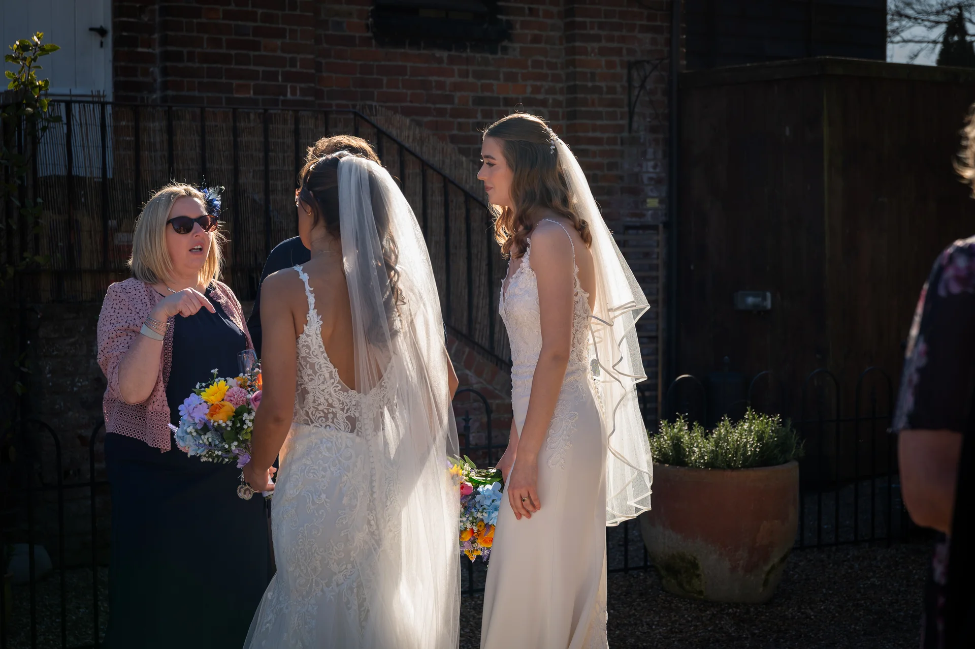 Two brides in wedding gowns and veils converse with a female guest outdoors in bright sunlight near a brick wall.