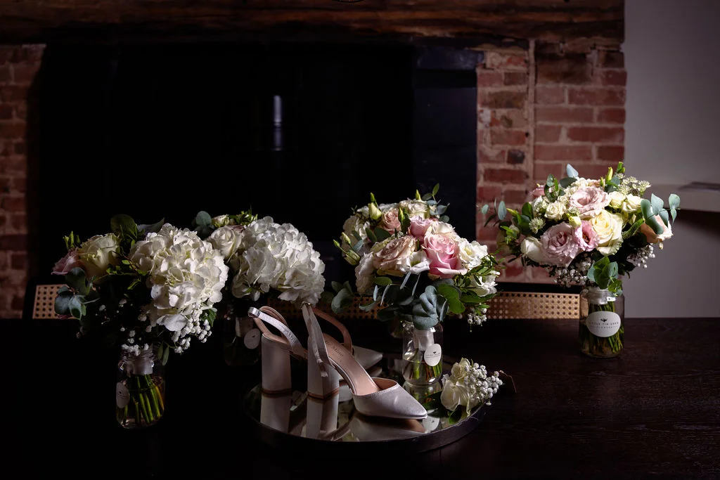 A bridal flat lay featuring white satin block heels resting on a mirror surrounded by floral arrangements in jars and a matching boutonniere against a rustic brick and fireplace backdrop.