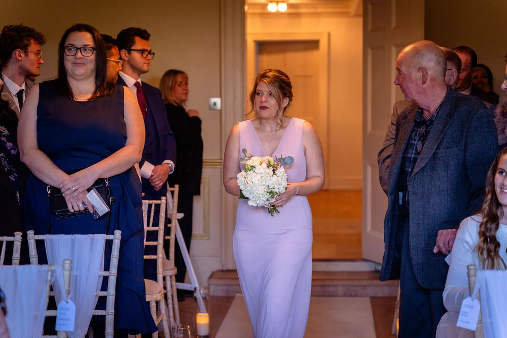 A bridesmaid walks down the aisle holding a white and green bouquet while guests watch from seated positions.