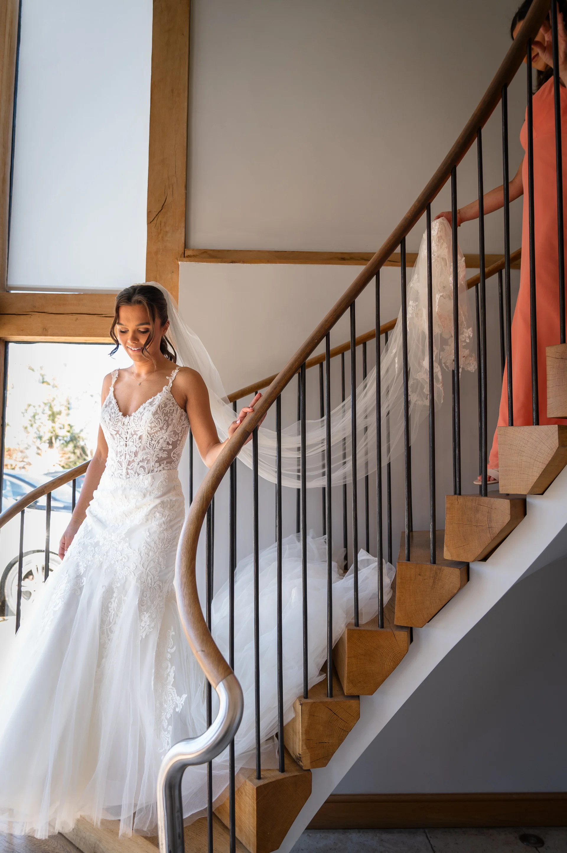 A smiling bride wearing an intricate lace a-line wedding dress descends a wooden spiral staircase while a bridesmaid holds her long veil from above.