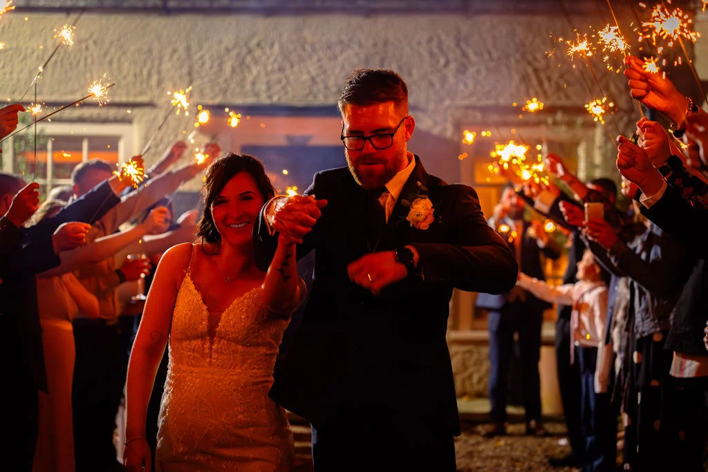 A newlywed couple happily walks hand in hand through a glowing tunnel of guests holding sparkling sparklers at night.