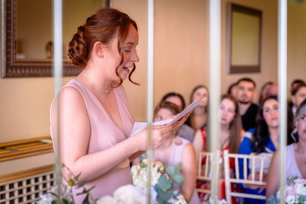 A bridesmaid in a pink dress reads her speech from a paper at the wedding breakfast table while seated guests look on.