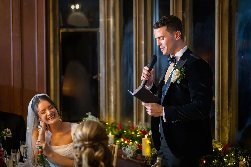 A groom in a black velvet tuxedo delivers a speech while holding a microphone, making the smiling bride laugh during the evening reception.