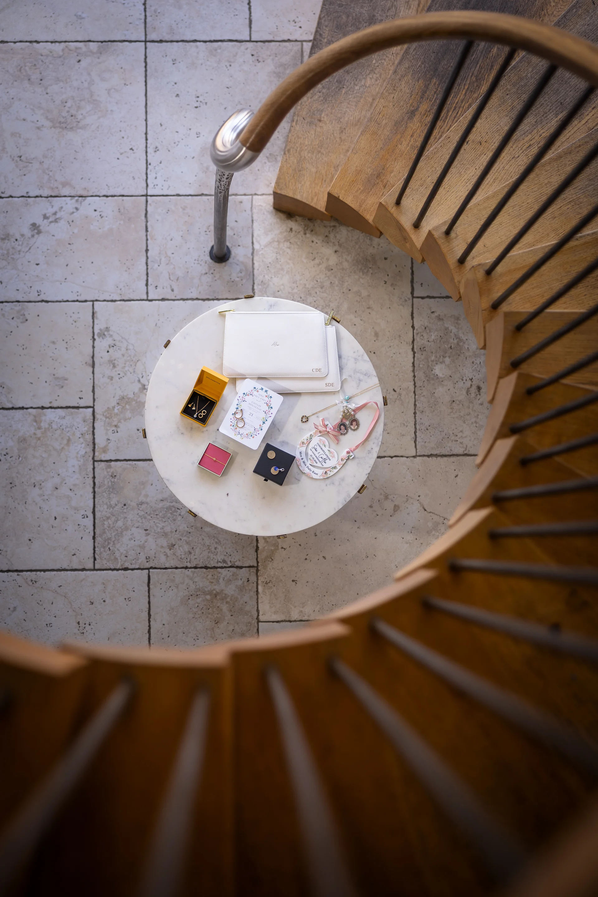 An overhead view looking down a wooden spiral staircase at a round marble table featuring a curated bridal flat lay with stationery, jewellery, and white leather clutches.