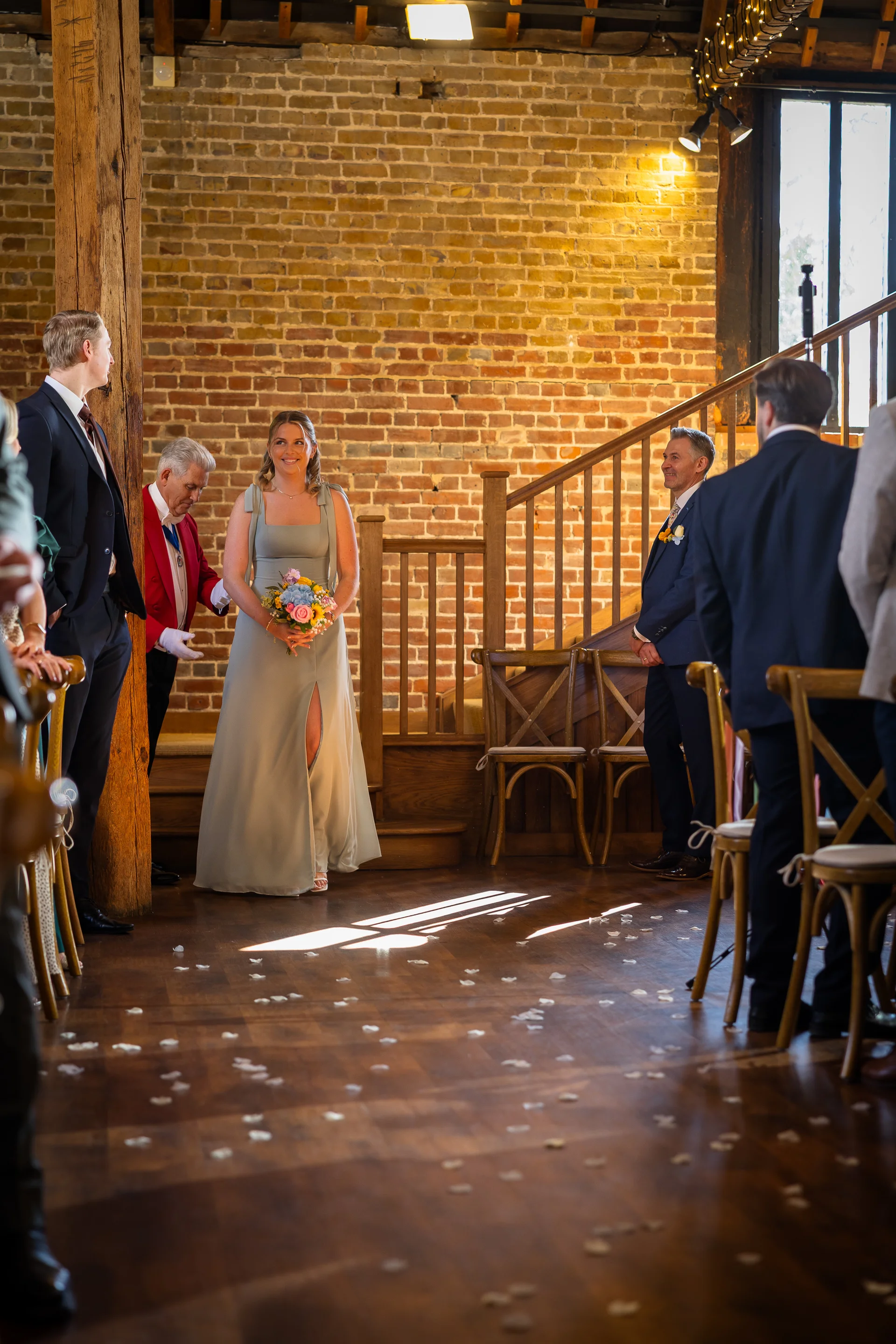 A smiling bride holding a colourful bouquet walks down the aisle in a rustic barn venue featuring exposed brick walls and a wooden staircase.