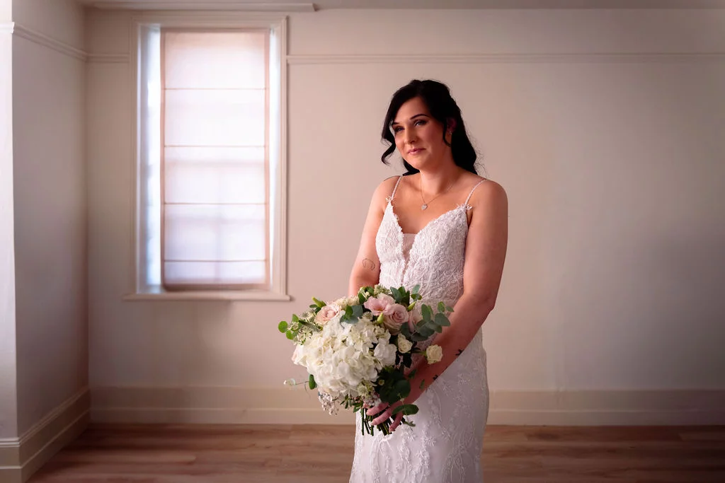 A stunning portrait of a bride standing gracefully near a window, illuminated by soft natural light while holding a lush hand-tied bouquet.