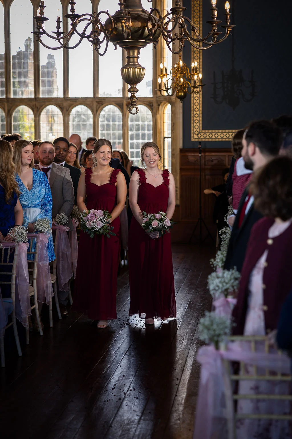 Two smiling bridesmaids in matching burgundy gowns walk down the wooden aisle of a historic hall decorated with hanging chandeliers and floral pew ends.