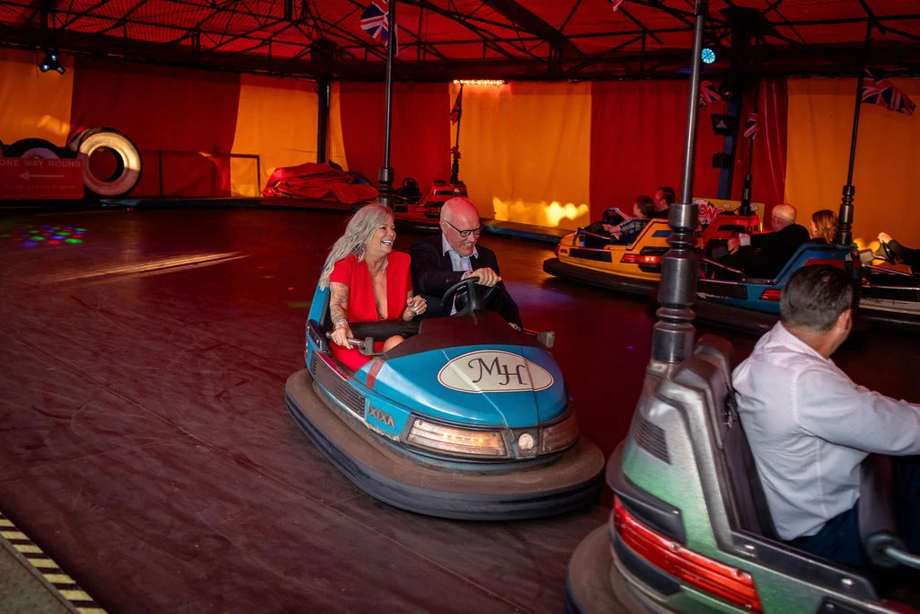 A joyful couple laughing while riding bumper cars together on the dance floor during an evening wedding reception celebration.