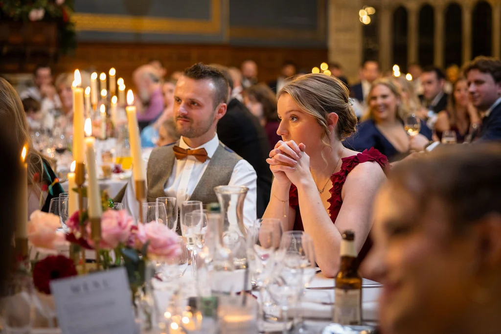 Wedding guests listen attentively during speeches at a candlelit banquet table decorated with pink roses and glowing taper candles.