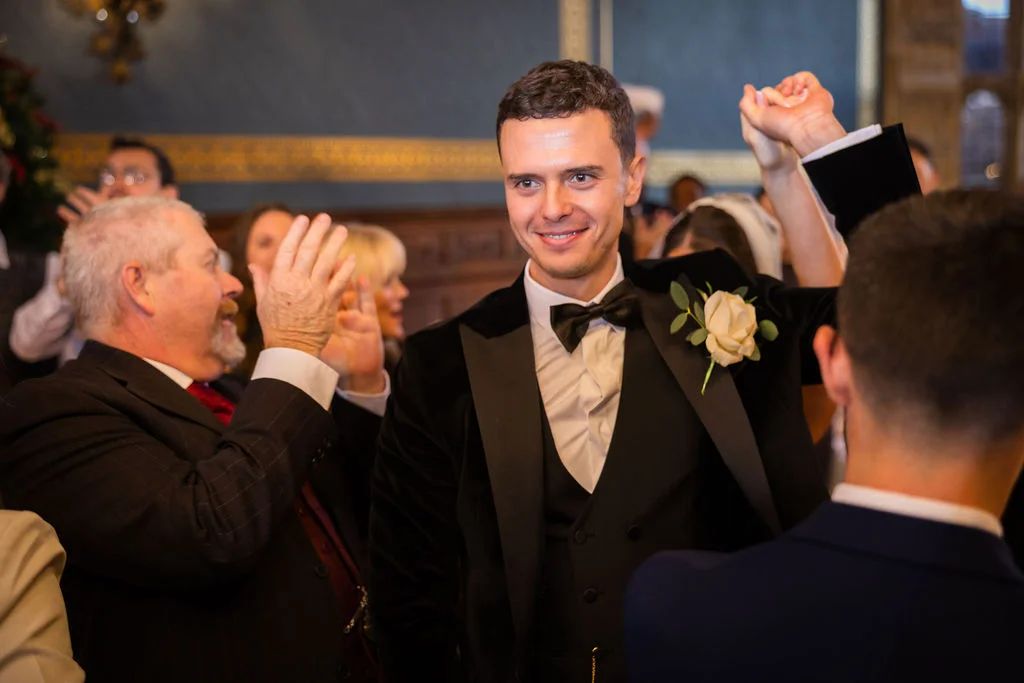 A smiling groom wearing a black velvet tuxedo with a white boutonniere looks towards the guests as they cheer and applaud during the wedding ceremony.