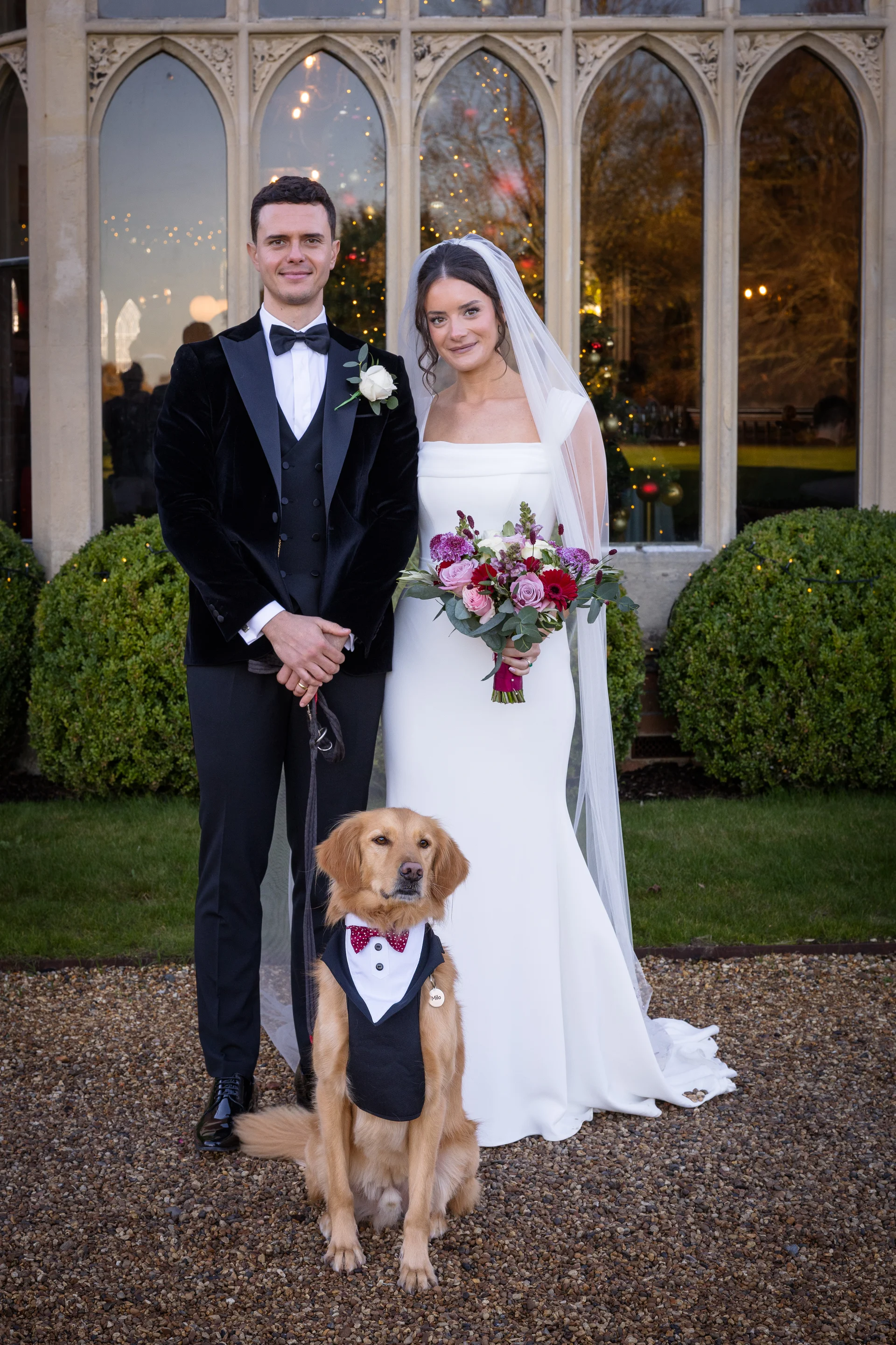 A newlywed couple poses outside a stately home with their golden retriever wearing a matching tuxedo and bow tie.