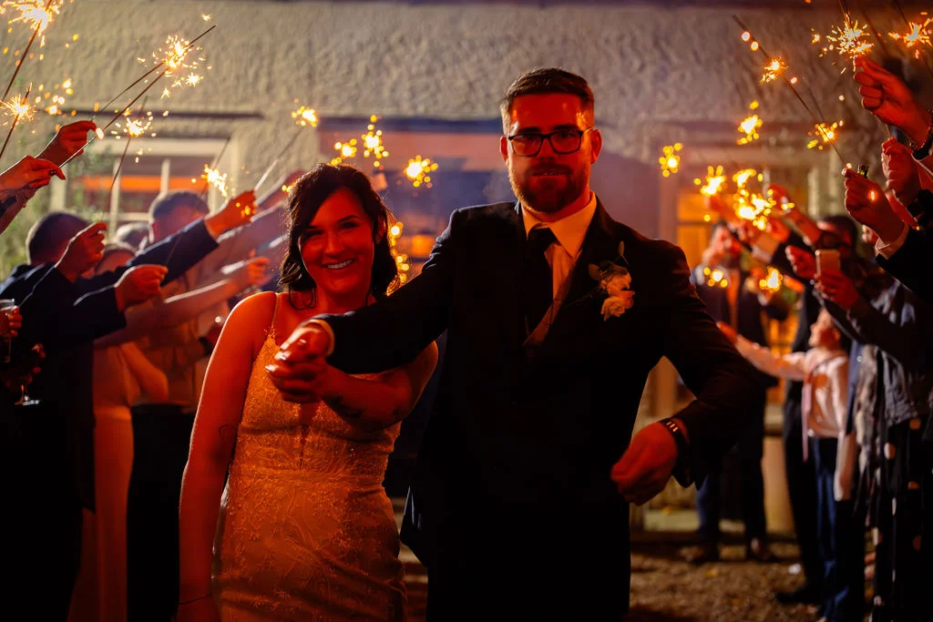 A smiling bride and groom walk hand in hand through an excited tunnel of guests holding glowing sparklers at night.