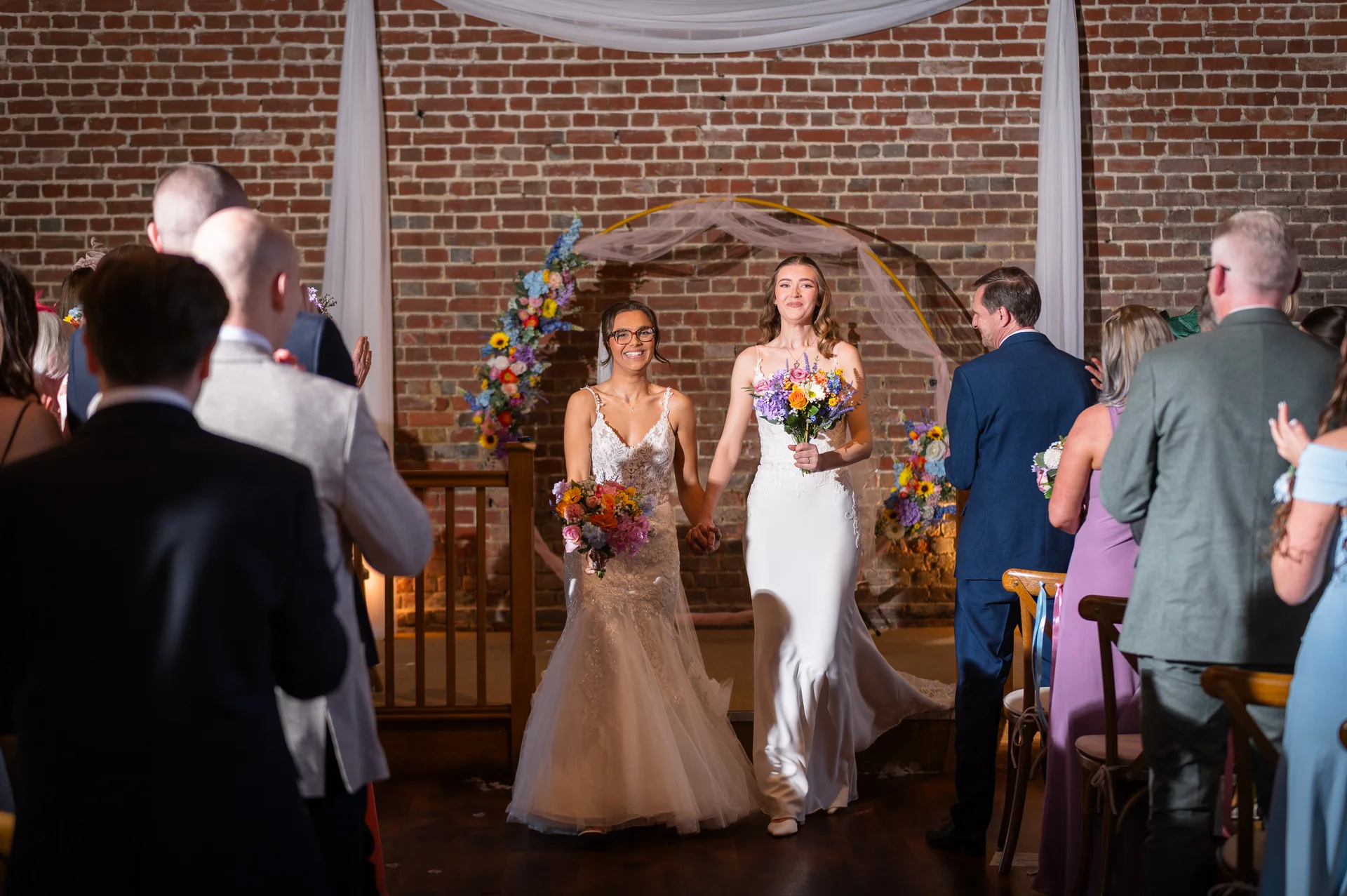 Two smiling brides walk down the aisle hand in hand, holding colorful bouquets, amidst cheering guests in an exposed brick barn ceremony venue.