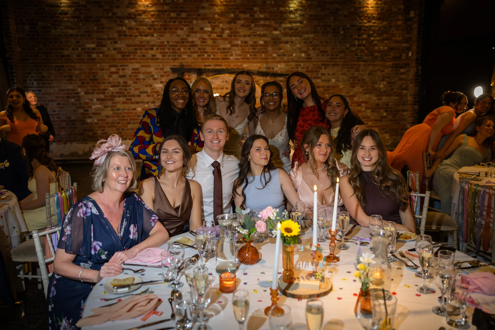 A smiling wedding party and guests gather around a candlelit round banquet table inside a rustic brick barn venue.