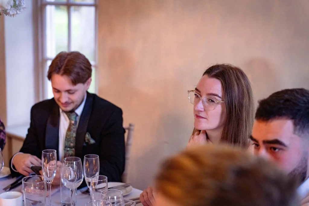 Wedding guests are seated together at a dining table during the wedding reception, illuminated by soft window light.