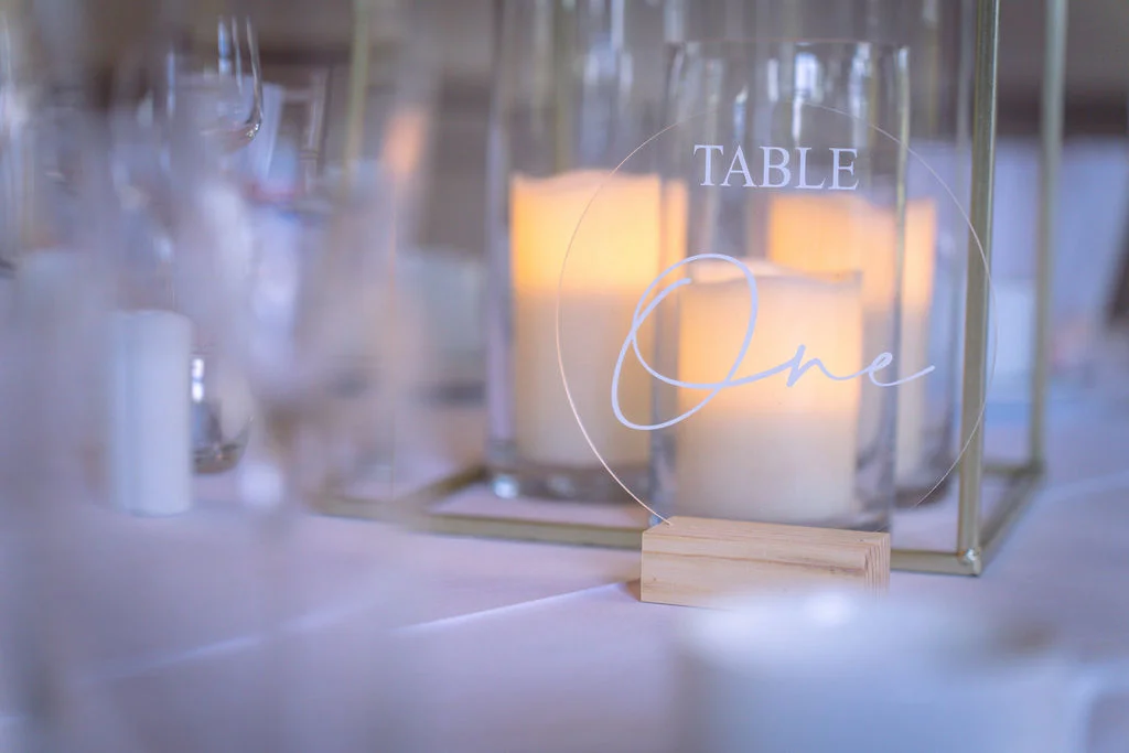 An elegant, minimalist table number sign made of clear acrylic displaying table one sits on a wedding reception table among glowing pillar candles and soft linens.
