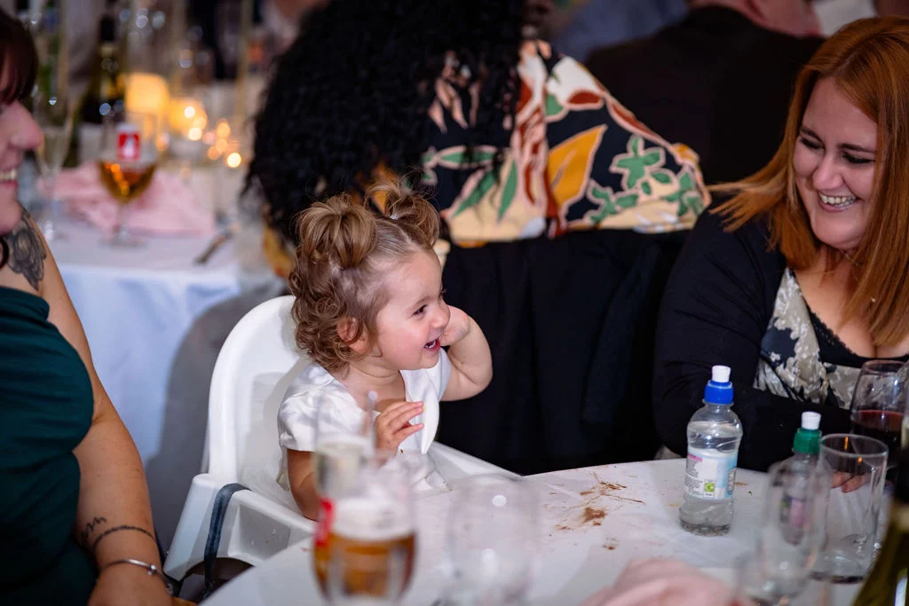 A smiling toddler sits in a high chair at a wedding reception table surrounded by joyful guests.