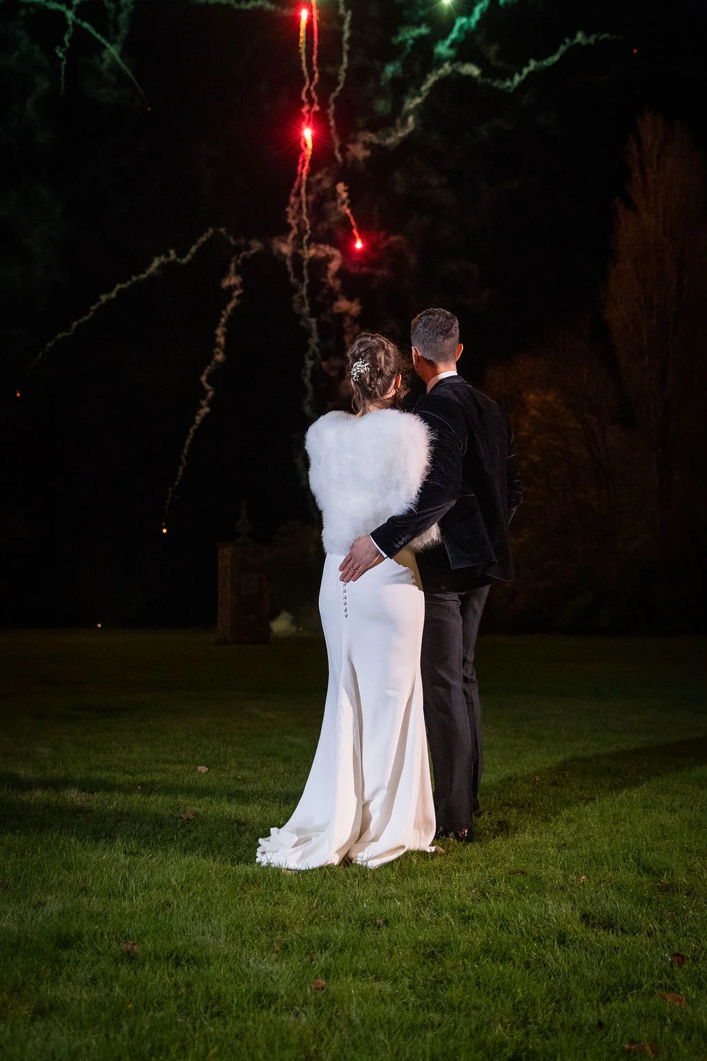 A newlywed couple stands on a lawn at night, wrapped in an embrace while watching a vibrant fireworks display light up the dark sky.