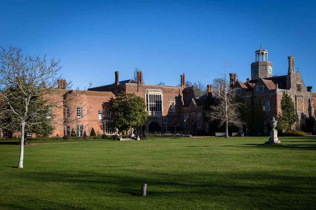 A wide exterior shot of a grand red brick country house wedding venue set against a clear blue sky with a sweeping lawn in the foreground.