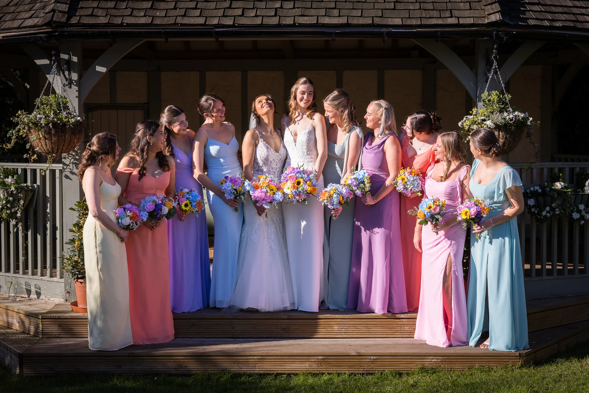 A joyful bride and her large group of bridesmaids wearing mismatched pastel dresses hold colourful bouquets while posing outside a wooden gazebo in the sunshine.
