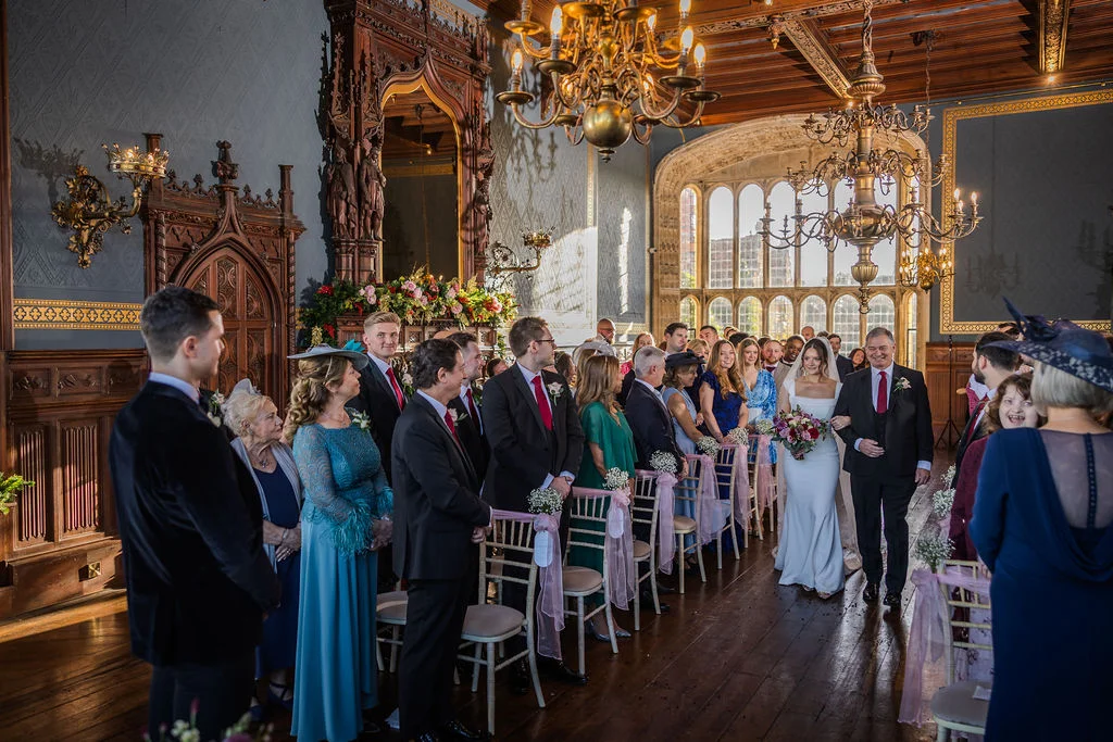 The bride walks down the aisle with her father inside a grand historic hall featuring large windows, wooden panelling, and chandeliers during the wedding ceremony.