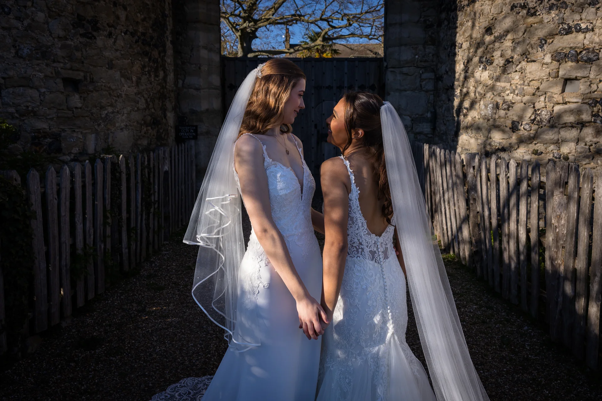 Two smiling brides in white wedding dresses with veils hold hands outside a historic stone gateway during golden hour.
