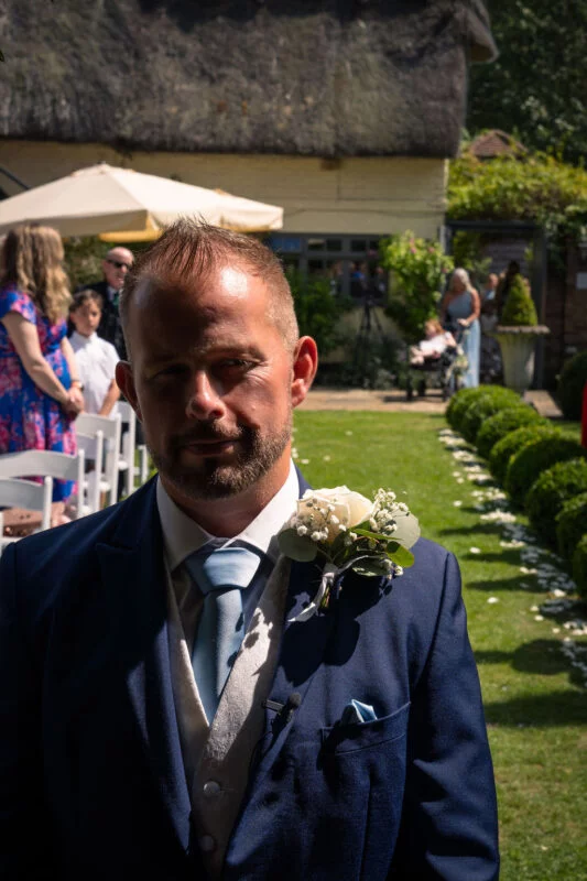 A groom in a navy suit and light blue tie stands outdoors in a garden with a white rose boutonniere pinned to his lapel.