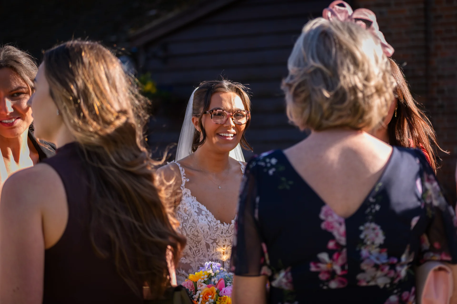 A smiling bride wearing glasses and a lace gown chats outdoors with wedding guests in the warm afternoon sunlight.