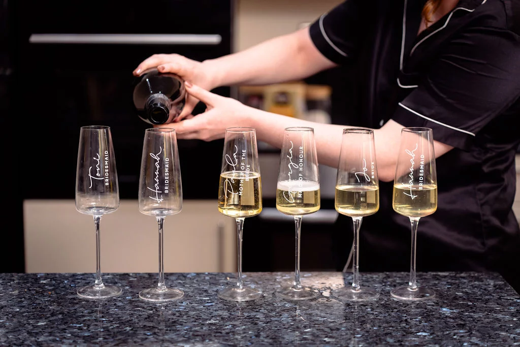 A person wearing matching pyjamas pours champagne into a line of personalised flutes resting on a granite countertop during bridal morning preparations.