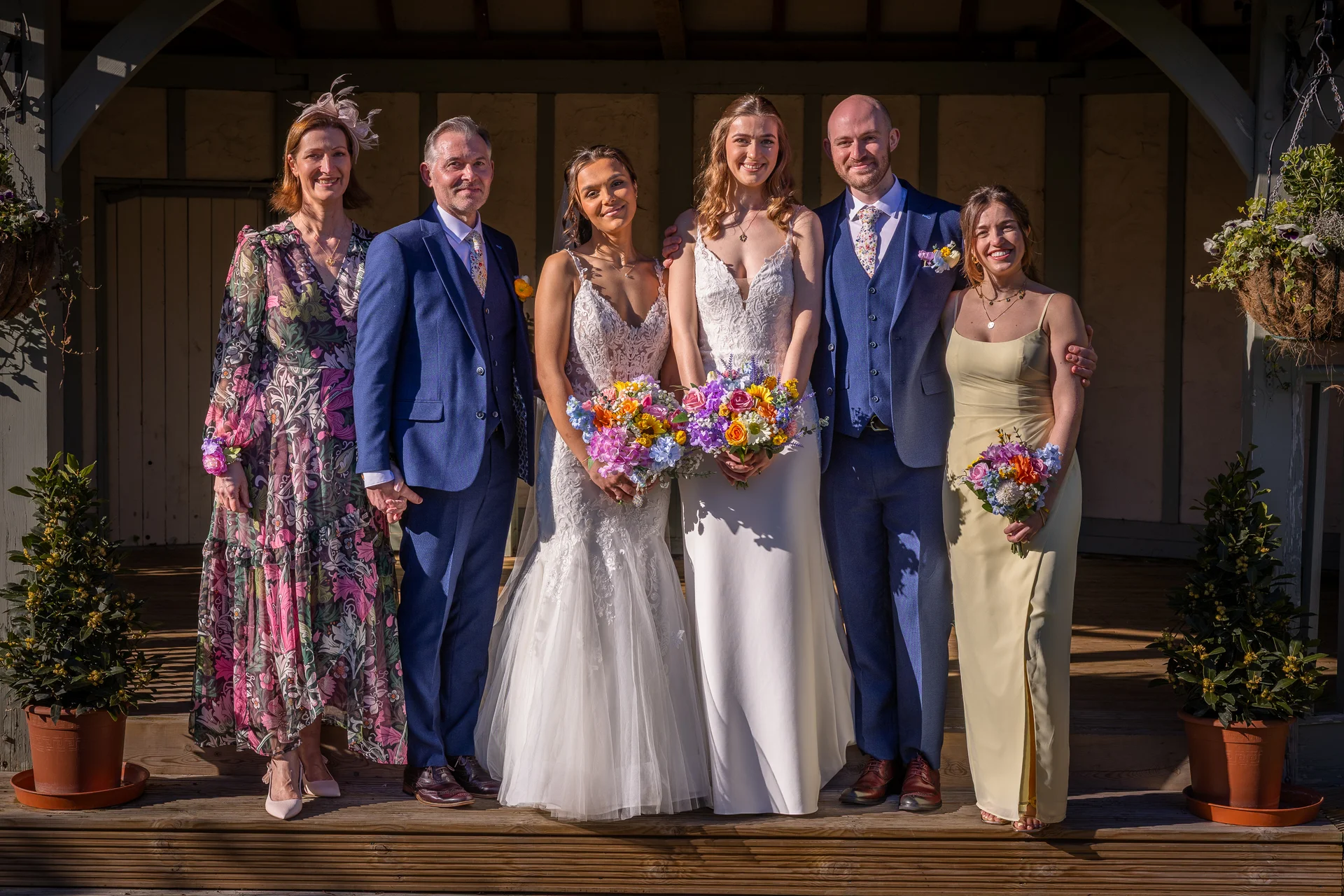 A joyous group portrait of two brides in wedding dresses flanked by family and wedding party members standing on wooden steps outdoors in bright sunlight.