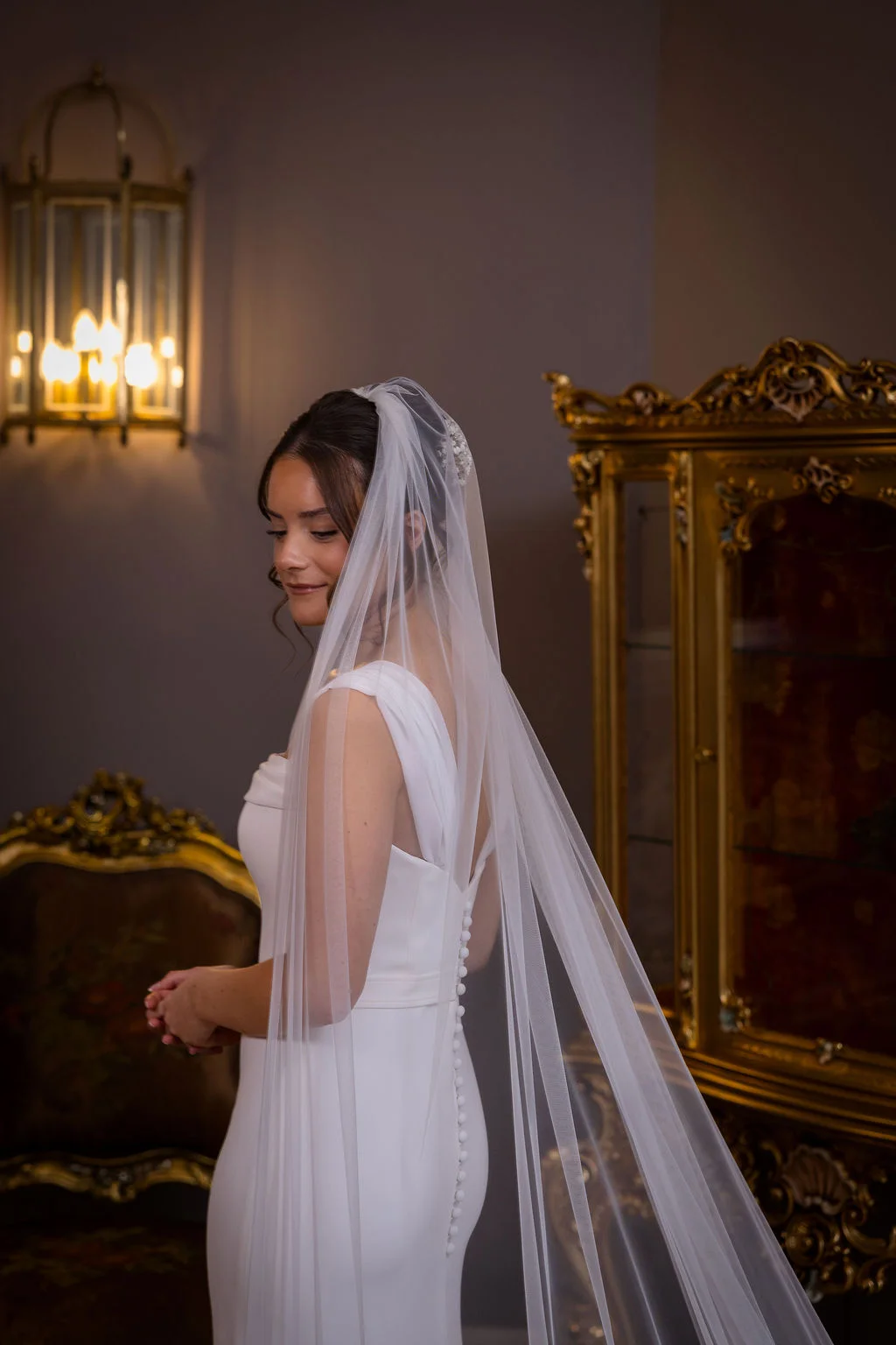 A stunning bride in an elegant white gown and cascading veil gazes downward softly inside an ornate room featuring antique gold furnishings and warm lighting.