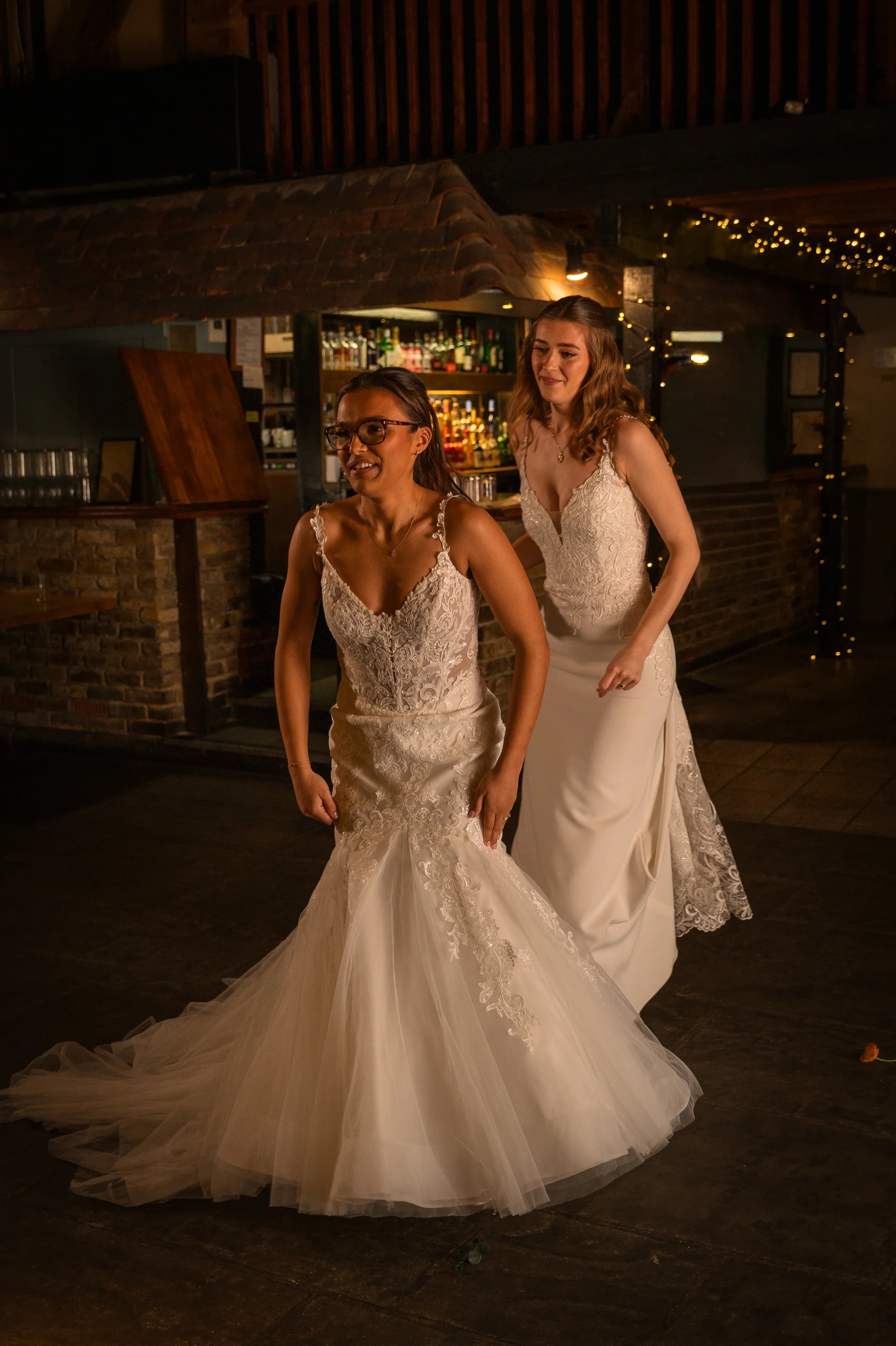 Two smiling brides in detailed lace wedding dresses dance joyfully together inside a rustic barn venue illuminated by string lights.