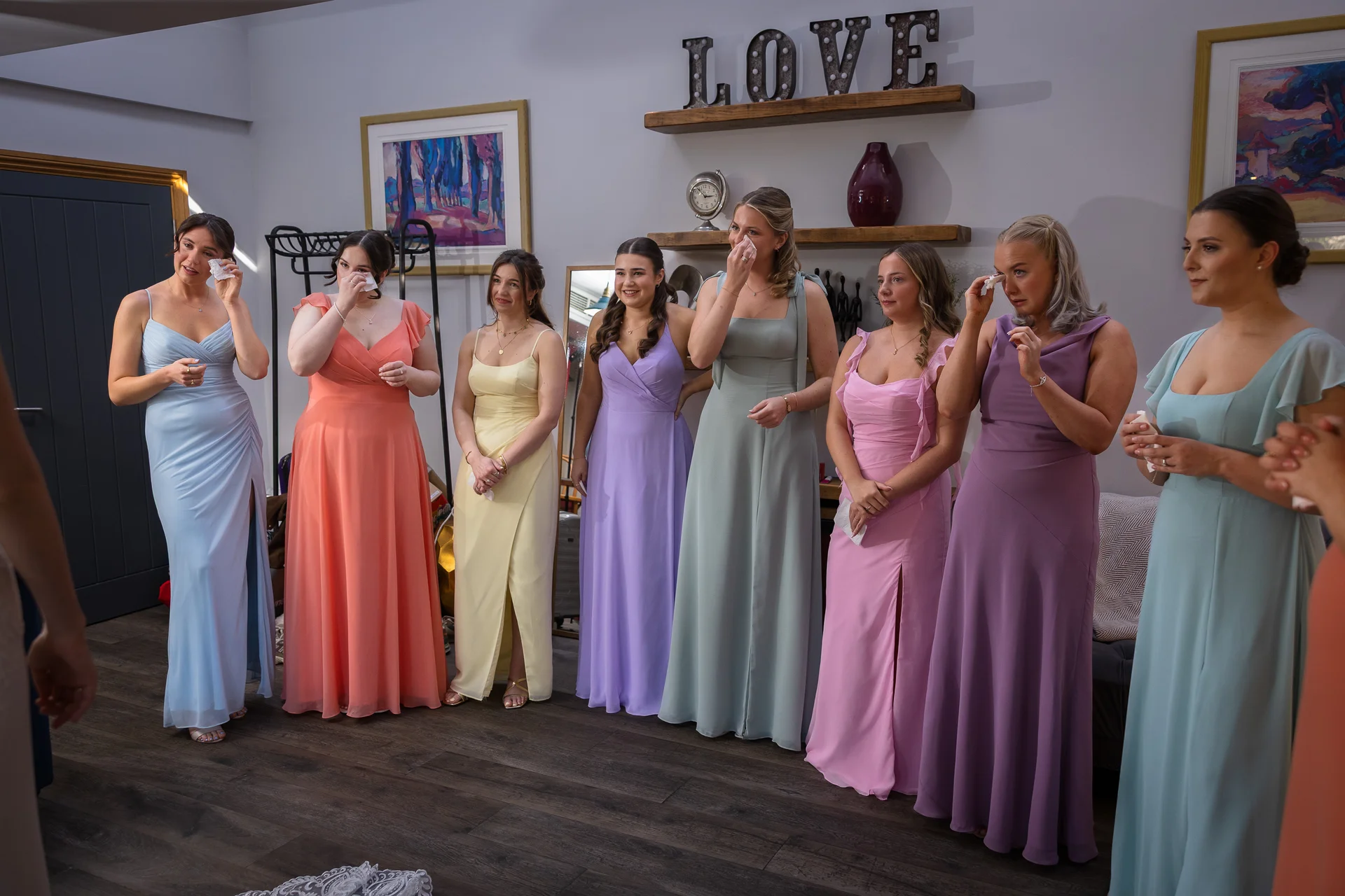 A line of emotional bridesmaids in colourful pastel dresses wipe away happy tears during a first look moment.