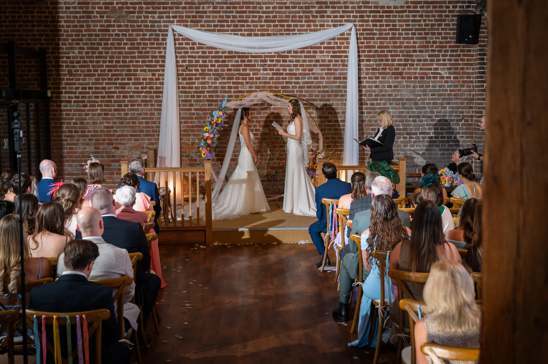 Two brides exchange vows during a rustic barn wedding ceremony framed by a floral arch and white fabric draping in front of seated guests.