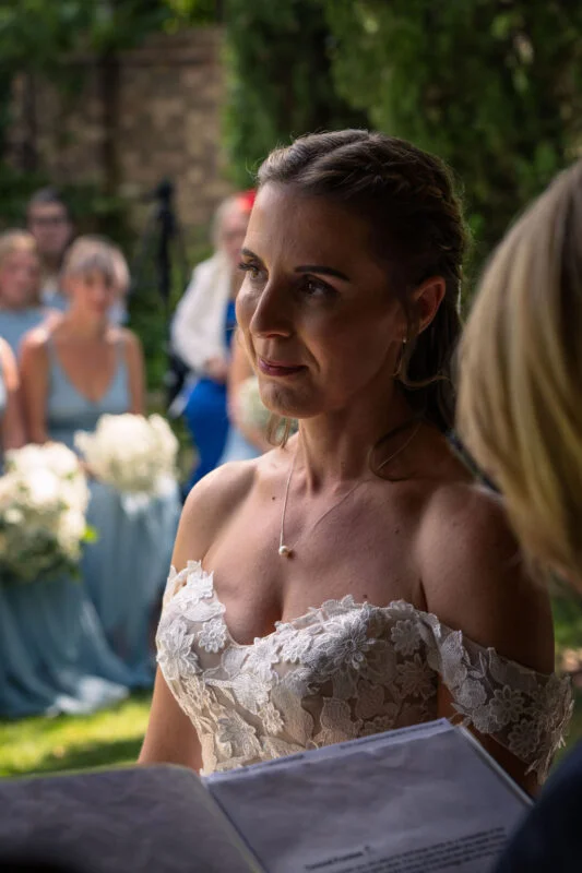 An emotional bride in a strapless floral lace wedding dress with an off-the-shoulder neckline listens intently during an outdoor wedding ceremony.
