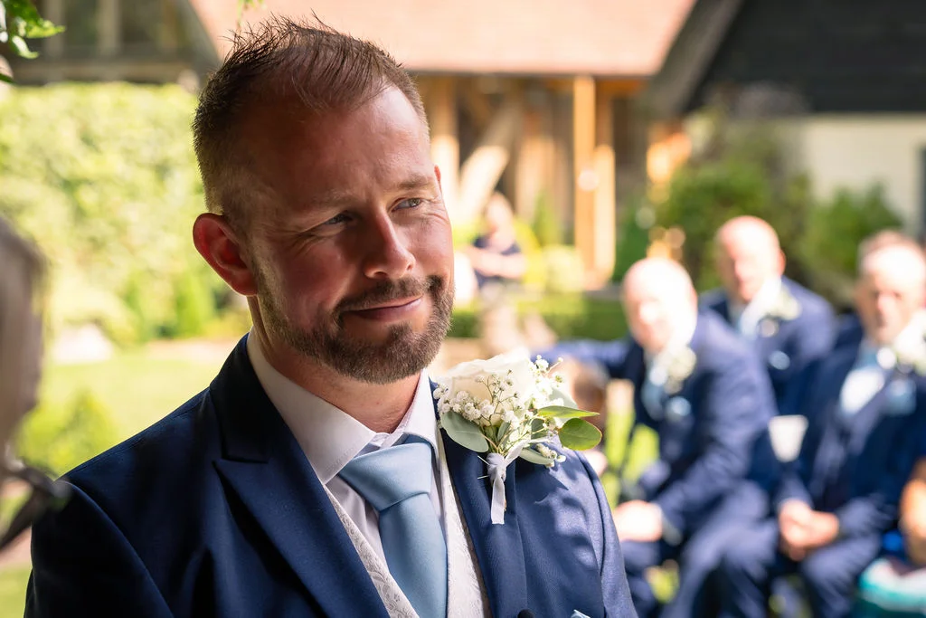 A groom wearing a navy suit and light blue tie smiles emotionally while waiting at the altar during an outdoor ceremony.