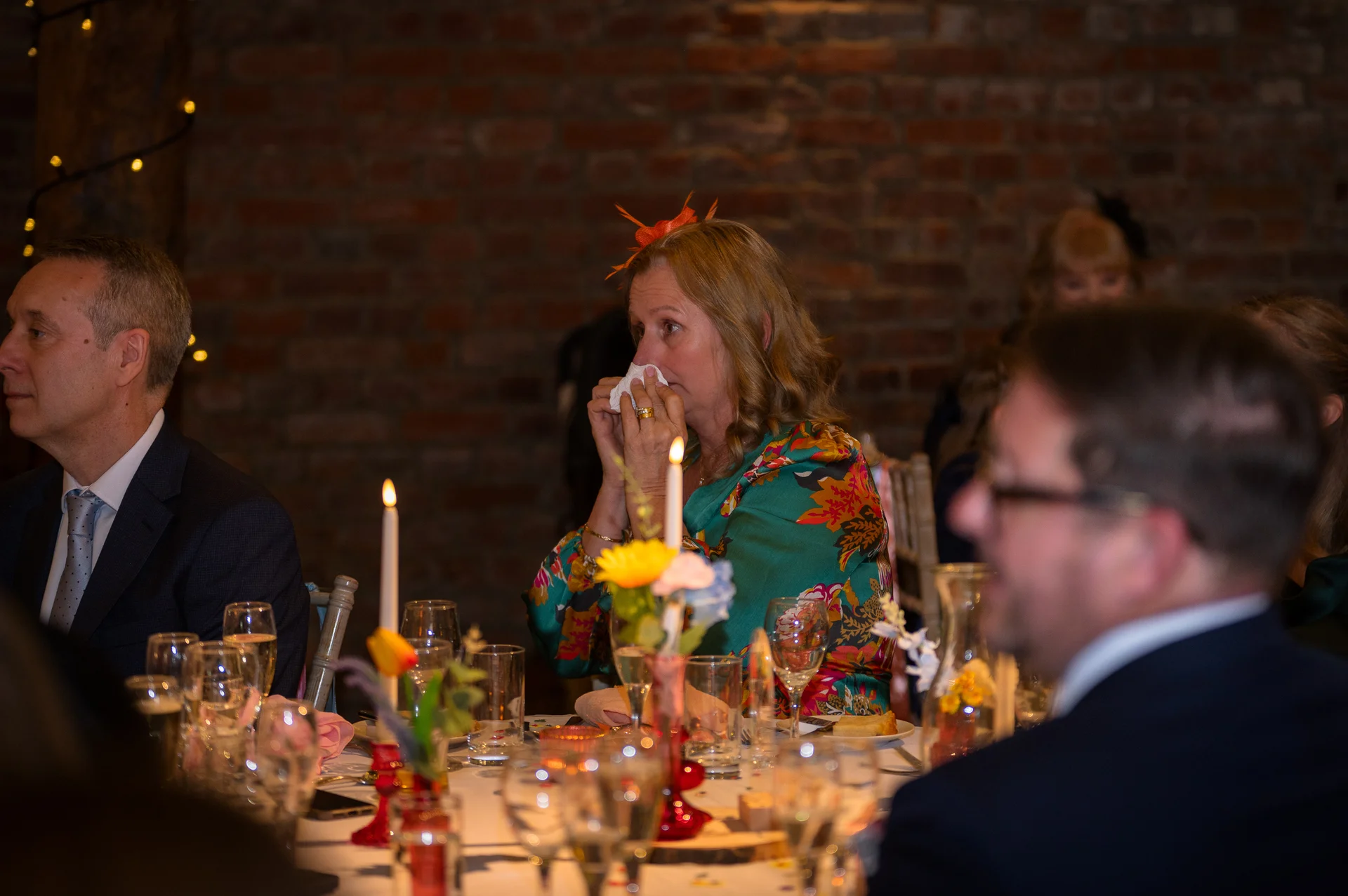 A female wedding guest sits at a candlelit reception dinner table, wiping a tear from her eye during emotional speeches while guests listen nearby.
